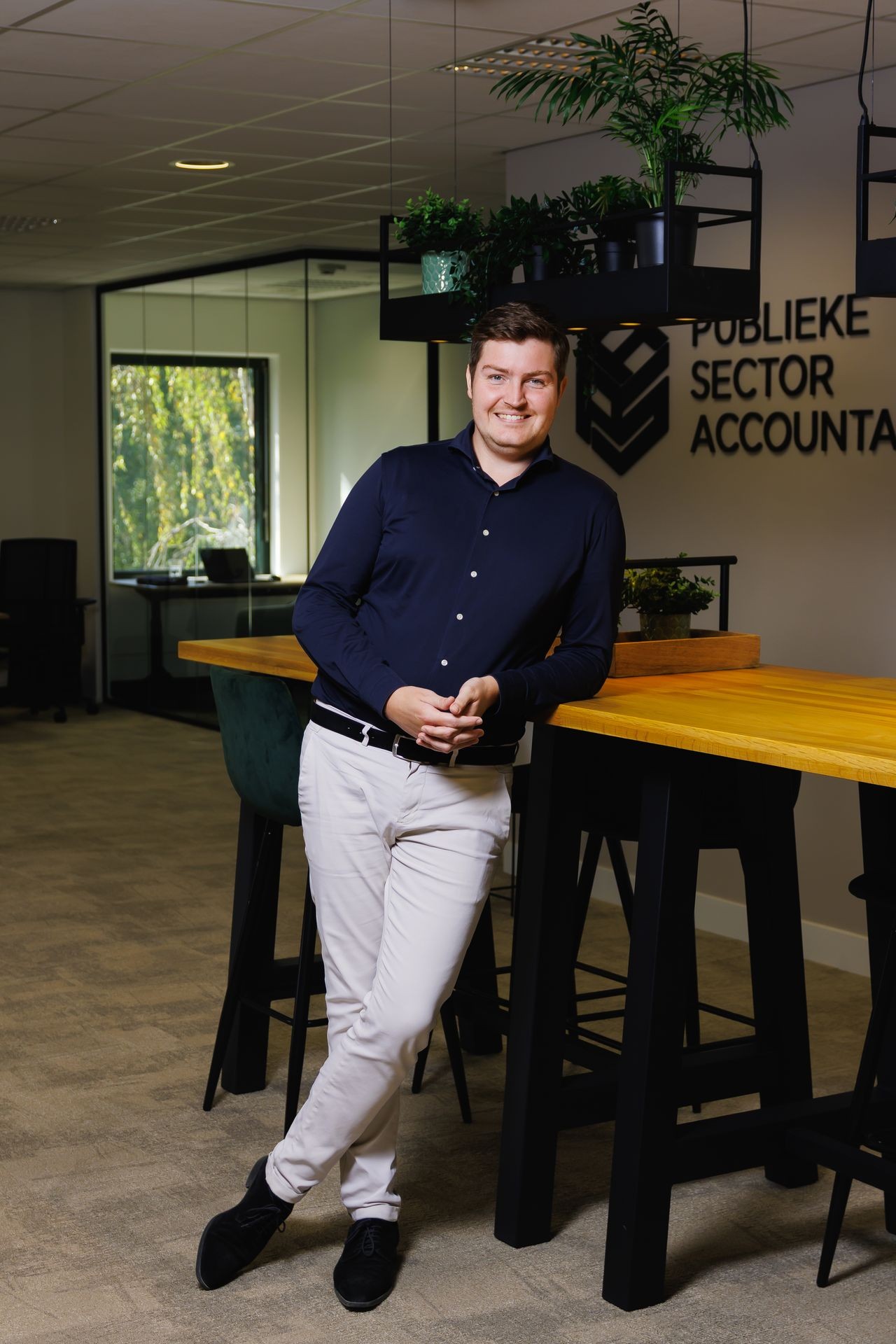 Collega die staat bij de hoge tafel in het kantoor van Publieke Sector Accountants, met groenplanten en bedrijfslogo op de achtergrond.