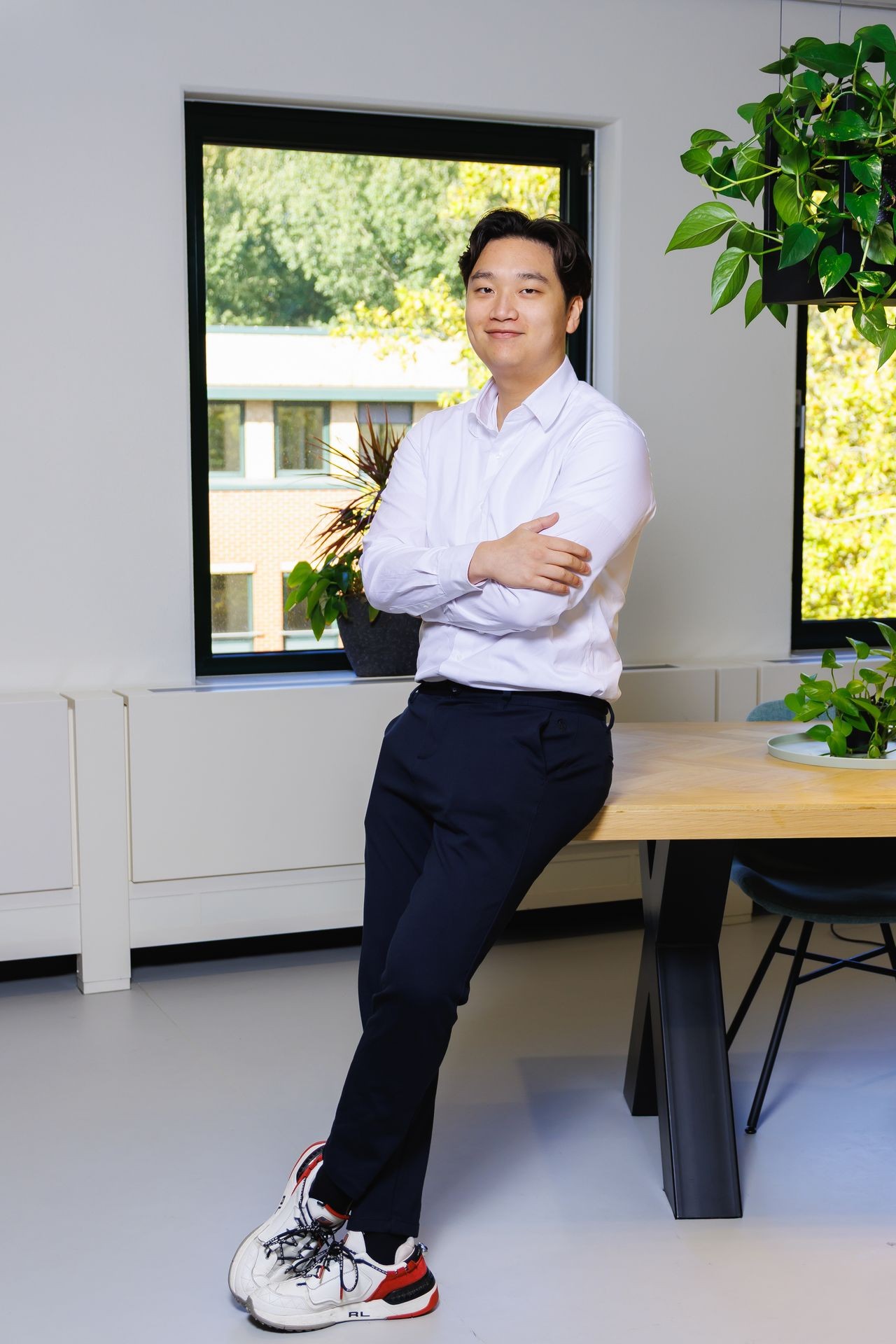 Collega die in een lichte kantoorruimte aan een bureau leunt, met planten en raam op de achtergrond.